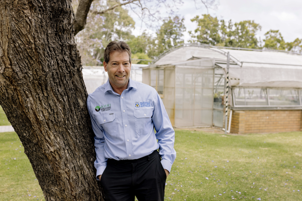 AHRI Director Hugh Beckie leaning on a tree in front of glass houses at UWA