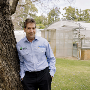 AHRI Director Hugh Beckie leaning on a tree in front of glass houses at UWA
